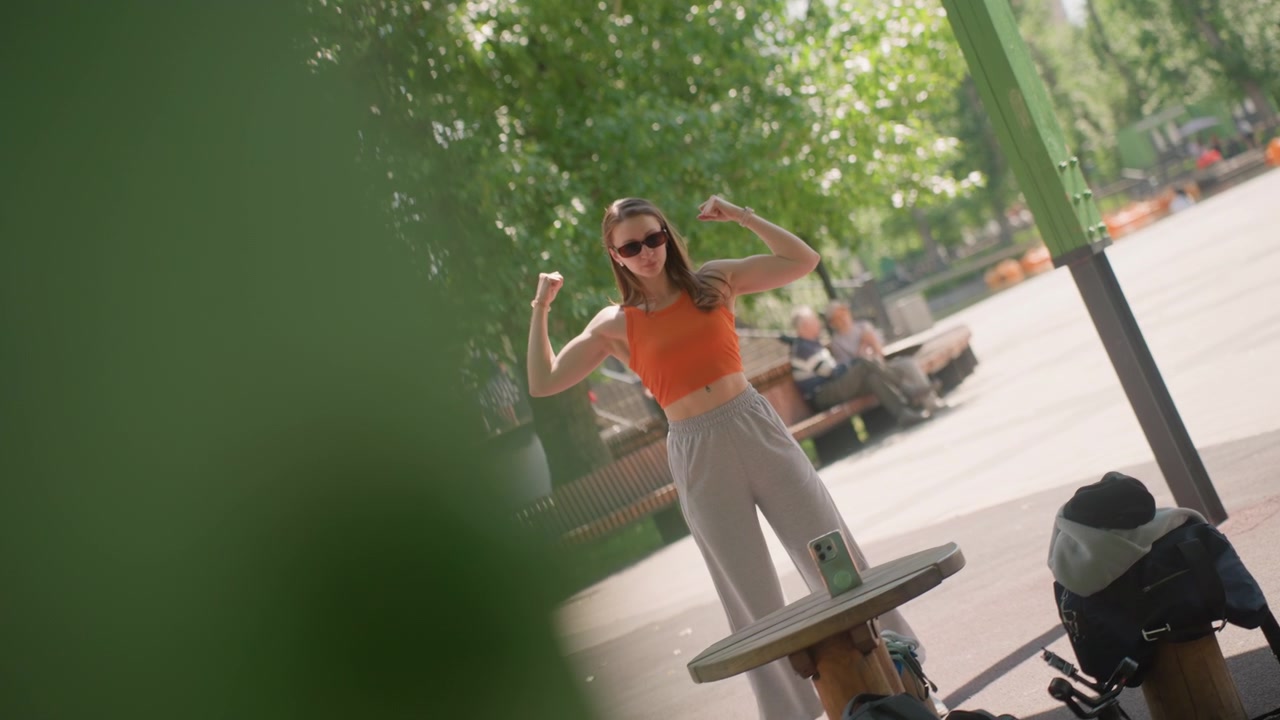 Giovane donna con crop top arancione e pantaloni grigi assume varie pose su un tavolo di legno all'aperto con alberi verdi e panchine del parco visibili. La donna flette le braccia mentre è in piedi accanto