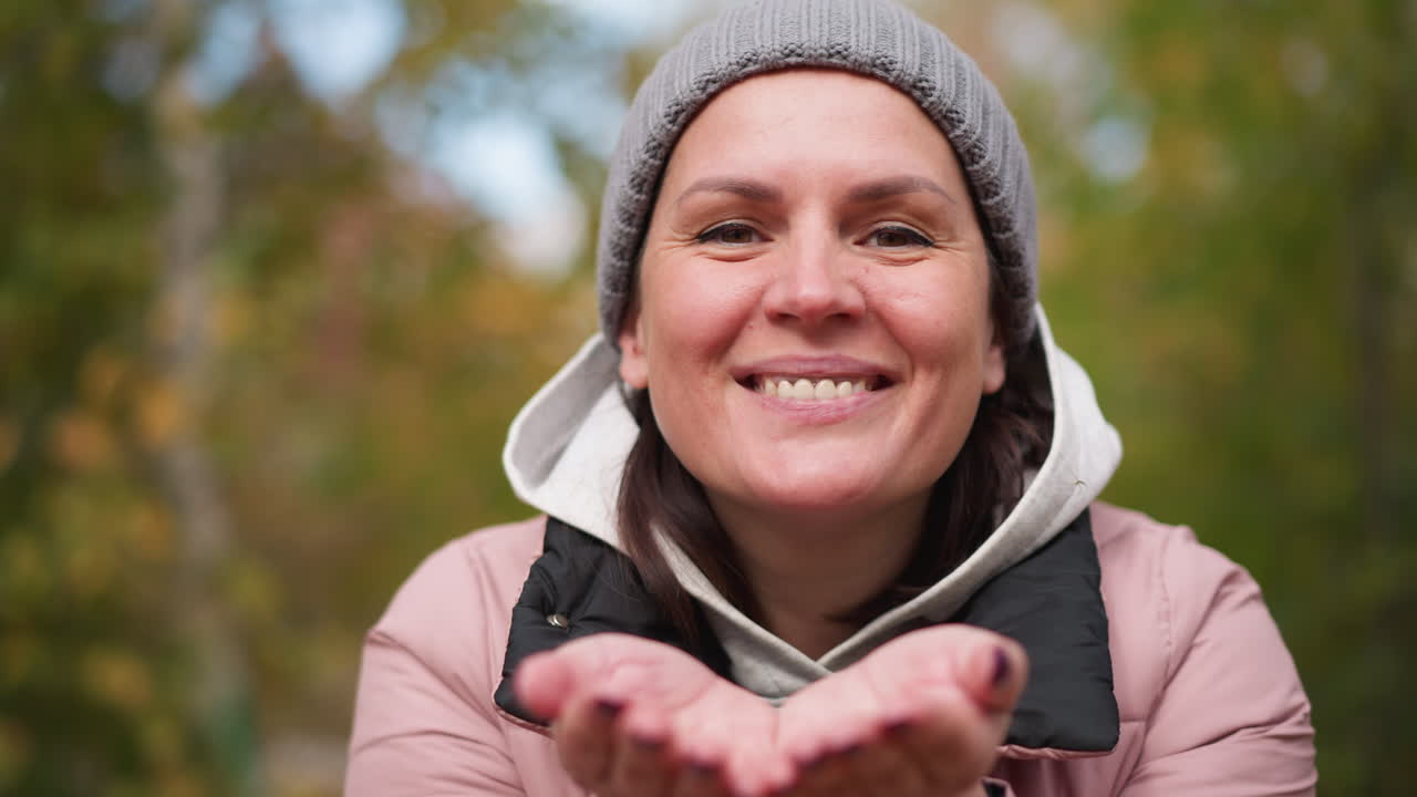 mujer con gorro de ceniza y chaqueta rosa soplando hojas secas de otoño de sus manos con una cálida sonrisa, uñas pintadas de negro, rodeada de colorido follaje borroso de otoño y árboles en el fondo