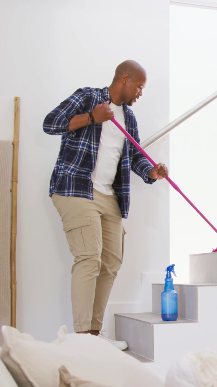 Vertical video: Holding pink mop, man in navy shirt cleaning stairs at home with blue sprayer