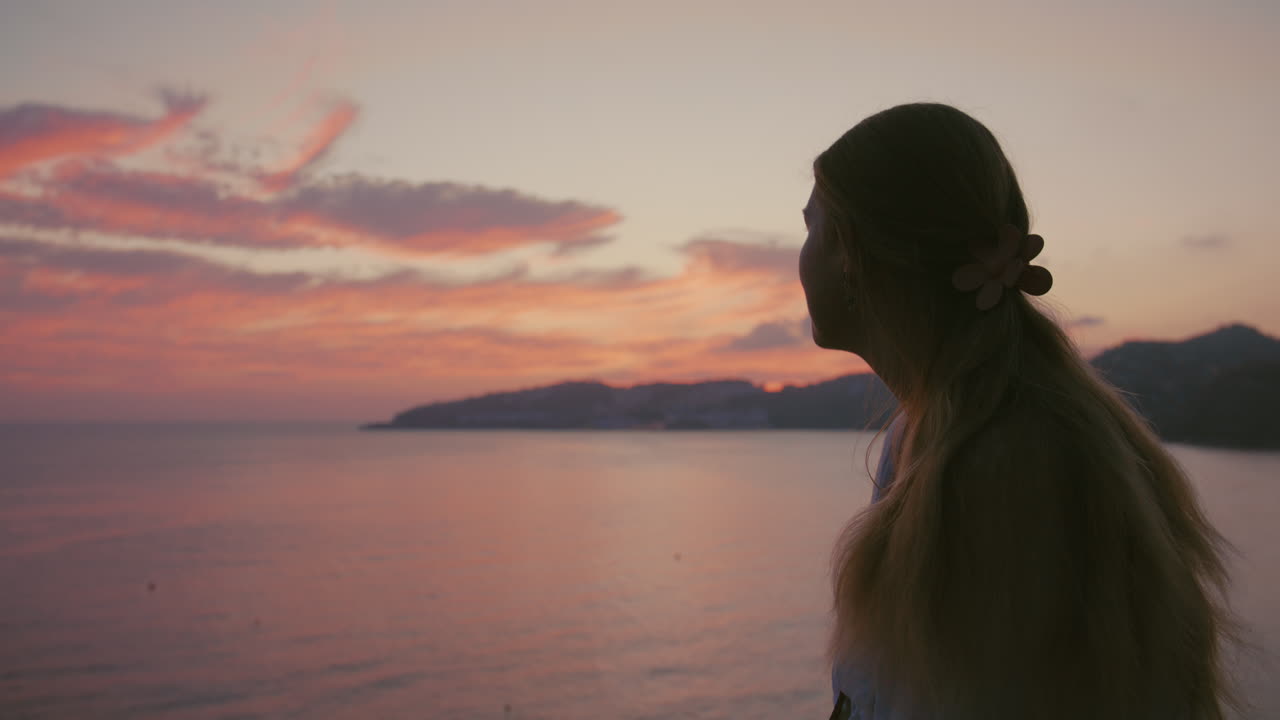 Side profile shot in soft silhouette, showing a young blonde woman gazing toward a vibrant, pastel-colored sunset. Peaceful, beautiful, and dreamy introspection
