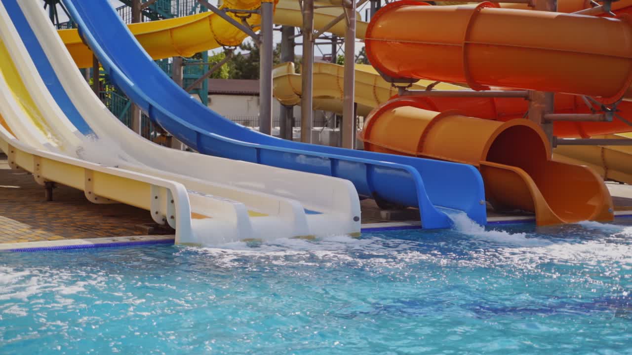 Colorful slides in the outdoor aqua park. Happy boy sliding down from slide into the swimming pool in summer. Slow motion.