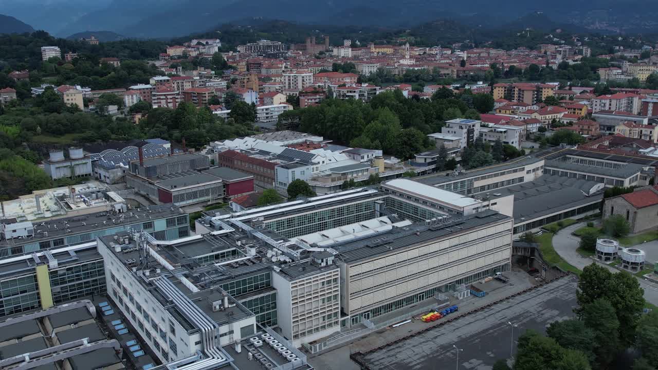 Modern glass-and-steel ICO IV building in Ivrea's Unesco-listed Olivetti district, framed by the Alps and Mount Mombarone in the background, drone shot, slow motion, sunny and cloudy sky