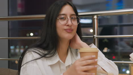woman in white shirt places hand on wooden table while sipping coffee with calm and warm smile, seated in chair near shiny metal railing and large glass window revealing blurred city night lights
