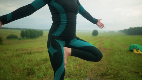 advanced woman stands with right leg in tree pose on colorful yoga mat, practicing yoga in a vast grassy field with trees in the distance and a yellow boot with green cloth nearby under a cloudy sky