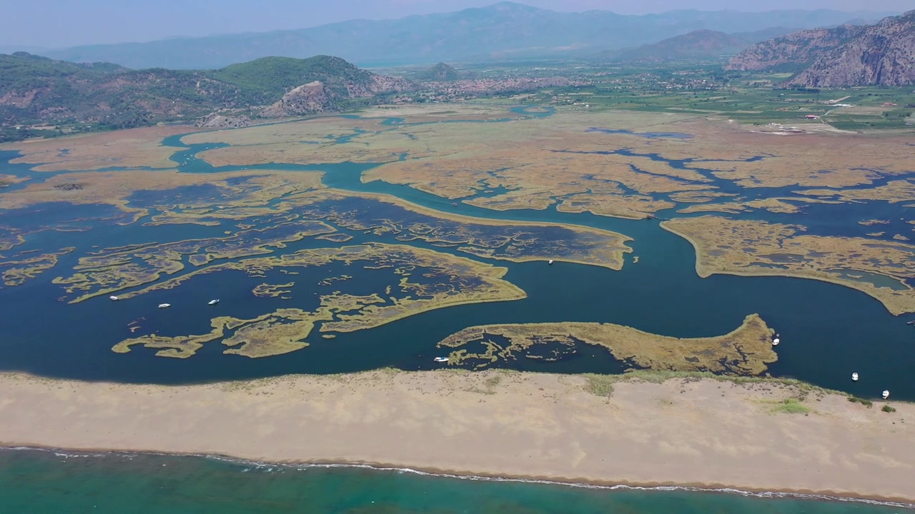 Muğla, Türkiye'de, Dalyan Nehri sazlık deltasının turkuaz Akdeniz ile buluştuğu İztuzu Plajı'nın yeşil dağlarla çevrili, açık ve güneşli bir günde yüksekten çekilmiş havadan drone panoraması.