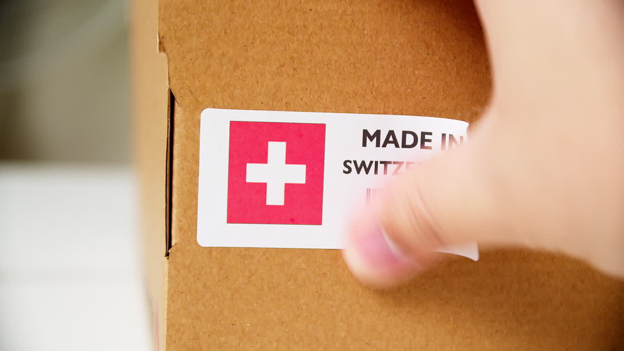 Hands applying MADE IN SWITZERLAND  flag label on a shipping cardboard box with products. Close up shot with premium quality barcode