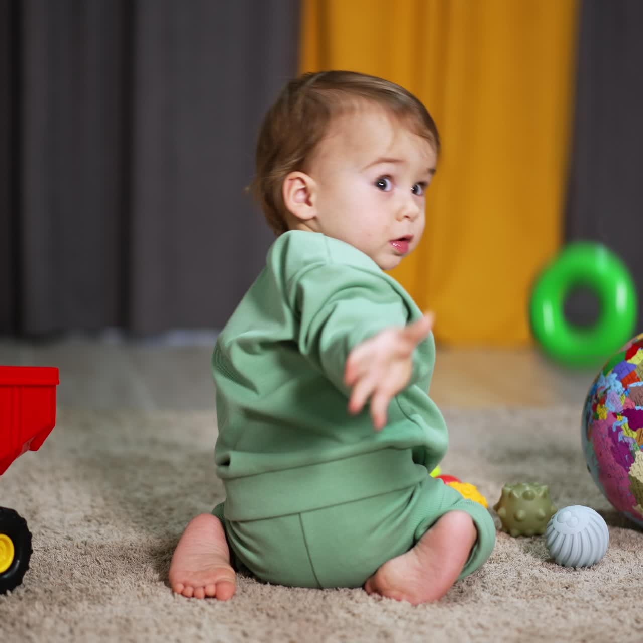 Active Caucasian toddler playing with toys on the floor indoors. Lovely kid takes a green plastic bagel and throws it into camera
