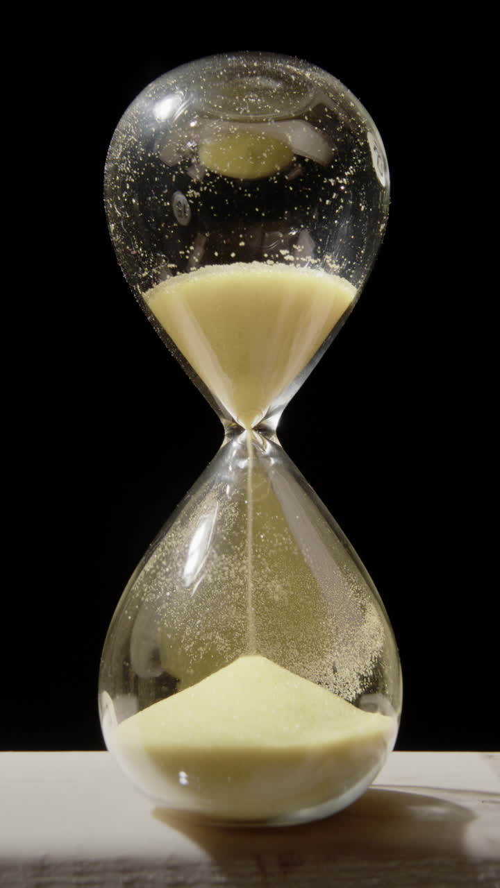Vertical shot of sand falling through hourglass bottle-neck, black background