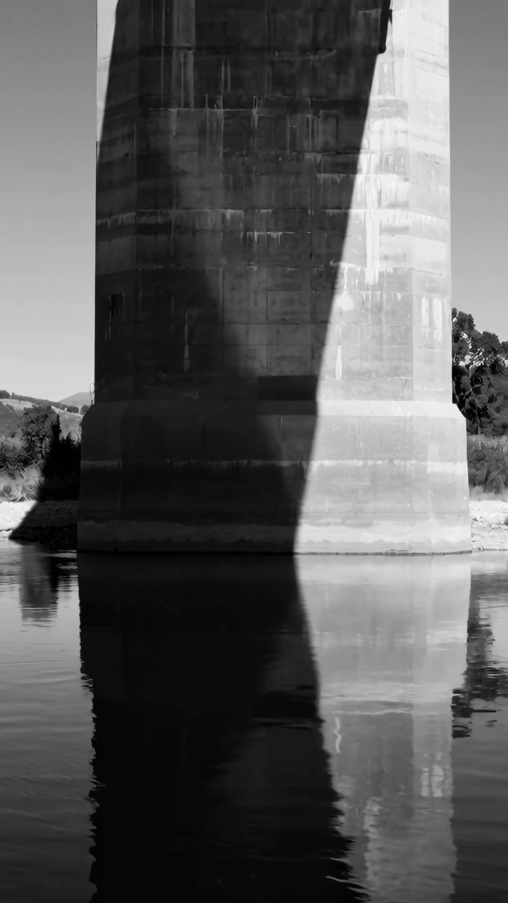 Vídeo vertical: Câmera inclinando-se revelando o topo do pilar de concreto de uma ponte refletindo uma cunha iluminada pelo sol na água