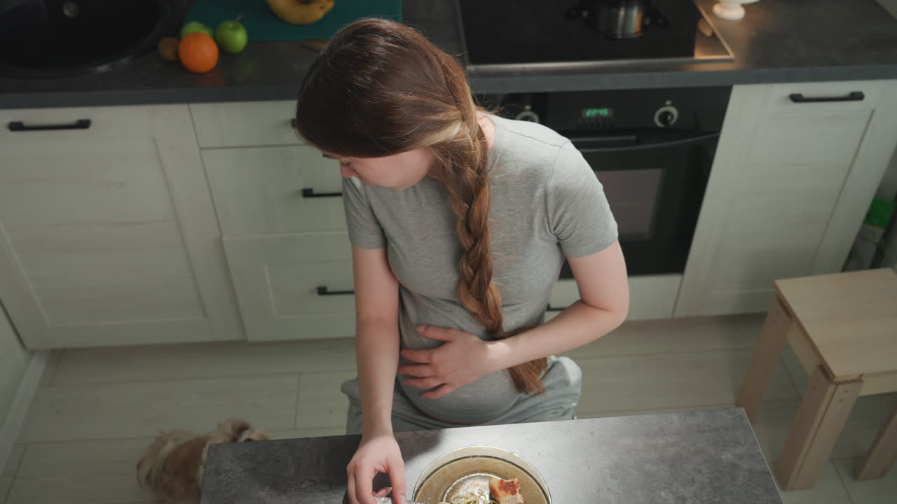 Pregnant caucasian woman in kitchen holds belly and reaches for bowl on counter, braided hair over shoulder, soft morning light, domestic calm, gentle craving and slight nausea while preparing small