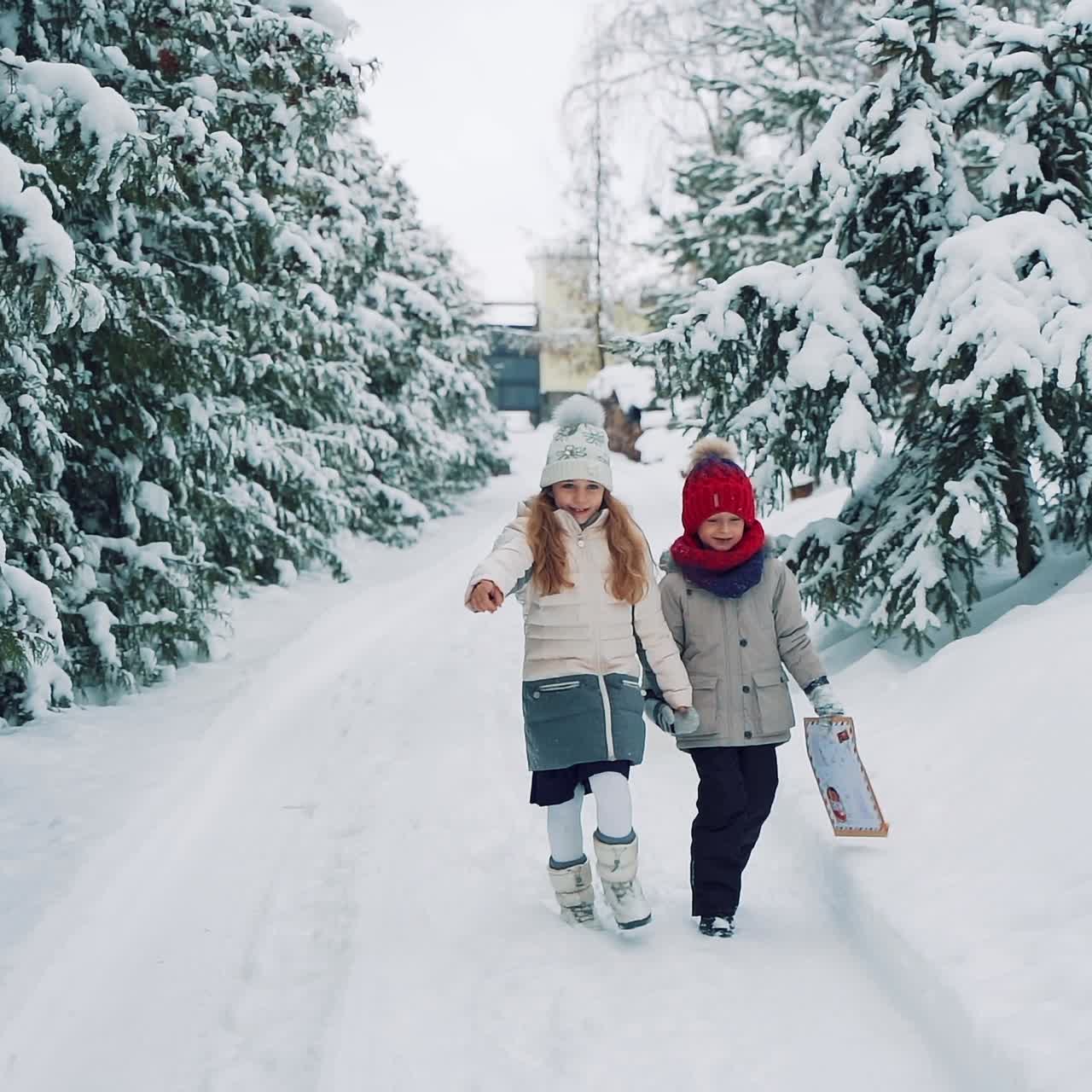 A little girl and a boy in warm clothes are walking along a snowy path holding each other's hands and talking on the background of green fir-trees. The boy is holding an envelope in his hands and listening to the story of a cute girl. Slow motion