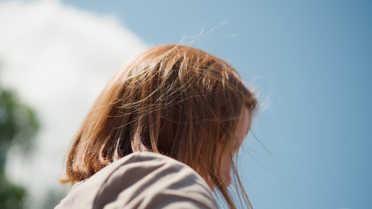Lady operating phone outdoors with gentle warmth on face, sunlight glowing through hair, serene atmosphere with blurred background and calm sky