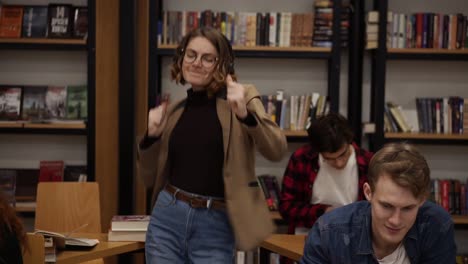 joyful excited european woman in jacket and jeans performing expressive dance while listening groovy music in headphones in academic library against bookshelves background and classmates around