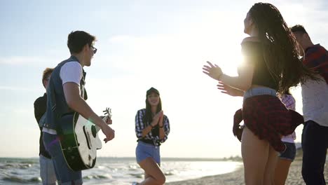 summer party on the beach. young friends drinking cocktails, dancing in the cirkle, playing guitar, singing songs and clapping on a beach at the water's edge during the sunset. slowmotion shot