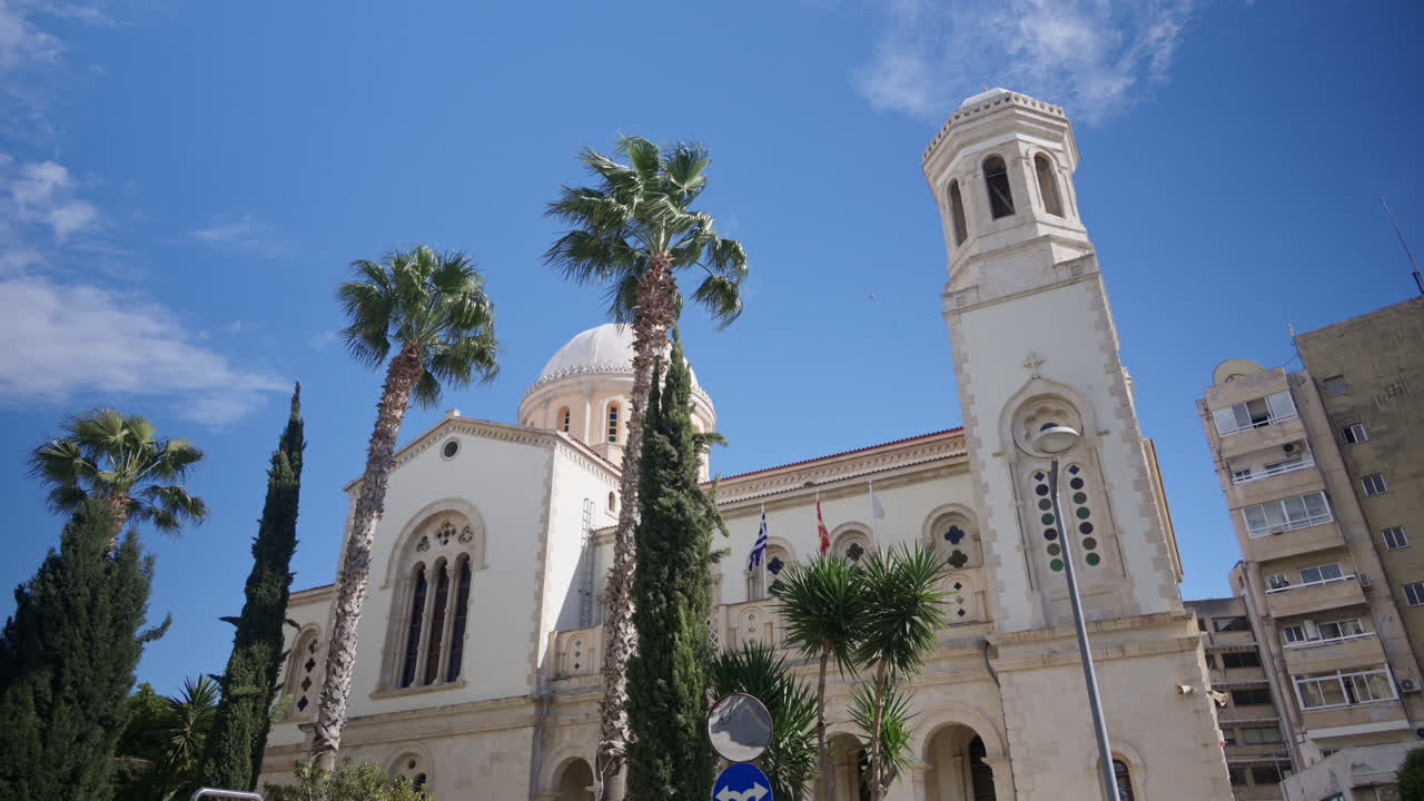 Vista em grande angular da Catedral de Ayia Napa em Limassol, Chipre, apresentando uma igreja ortodoxa tradicional com uma cúpula central e torre sineira rodeada por palmeiras sob um céu azul claro.