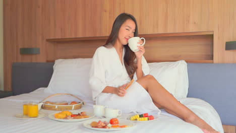 Hermosa-Mujer-Sentada-En-La-Cama-Tomando-El-Desayuno,-Tomando-Tostadas-Y-Oliendo-El-Café,-Habitación-De-Hotel