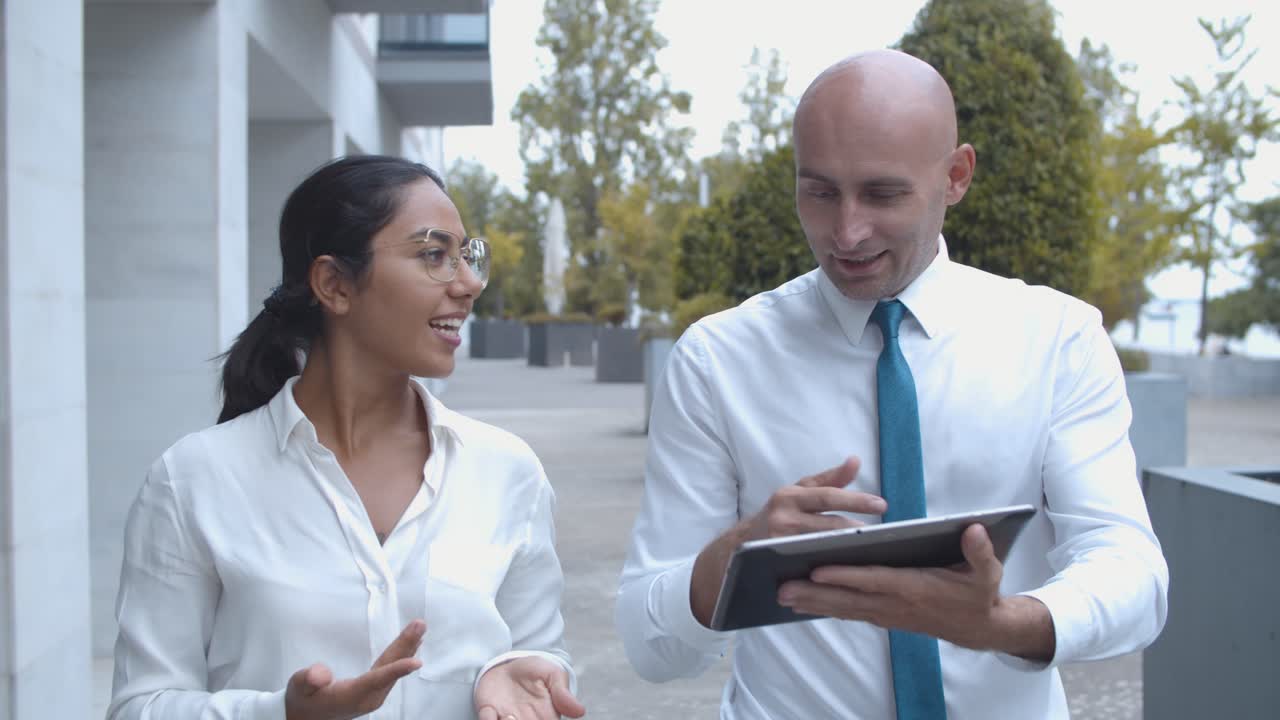 Front view of two business colleagues discussing project and watching presentation on tablet while walking at office building