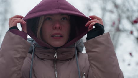 young lady shivering from cold pulls hoodie over head in chilly winter weather, reddened face and focused expression as she adjusts hood for warmth, blurred frosty background with bare tree branches