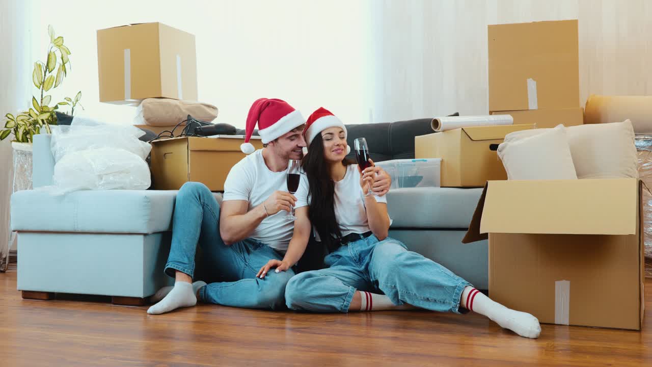 Young couple move into new apartment. New year period or Christmas time. Couple sit on floor and celebrate holidays in new apartment. Drink wine and have fun.