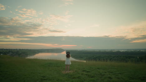 a woman in a white dress and hat stands thoughtfully in a grassy field overlooking a tranquil lake at sunset. she gazes into the distance, capturing a peaceful, reflective moment as the sun sets