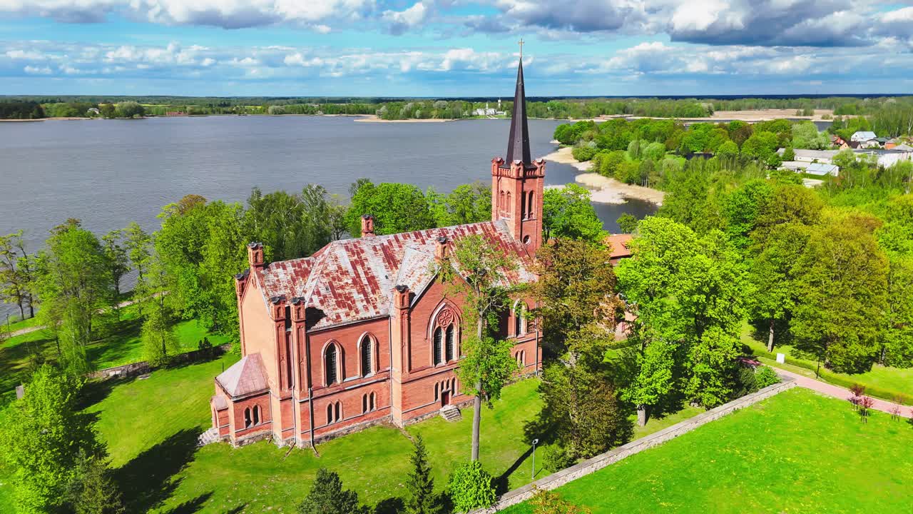 A historic red church stands beside a lake, surrounded by lush spring trees and green open fields, with a distant town and dramatic clouds on the horizon.