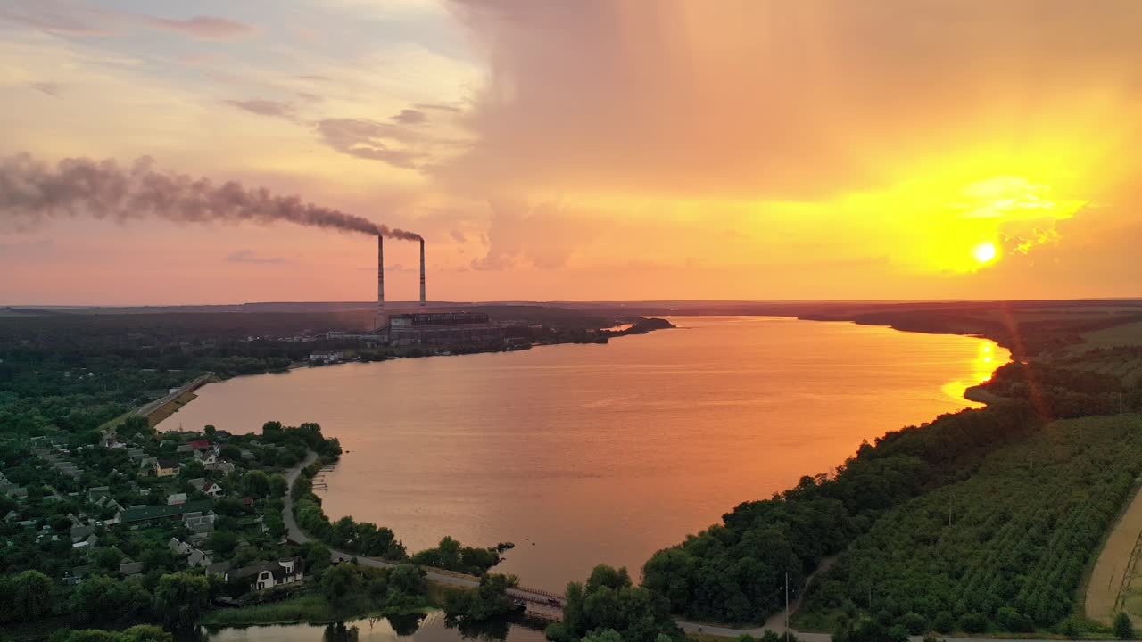 Rural background near the lake at sunset. Aerial view of pipes with smoke at the bank of the river in the evening. Amazing sunset over the beautiful scenery.
