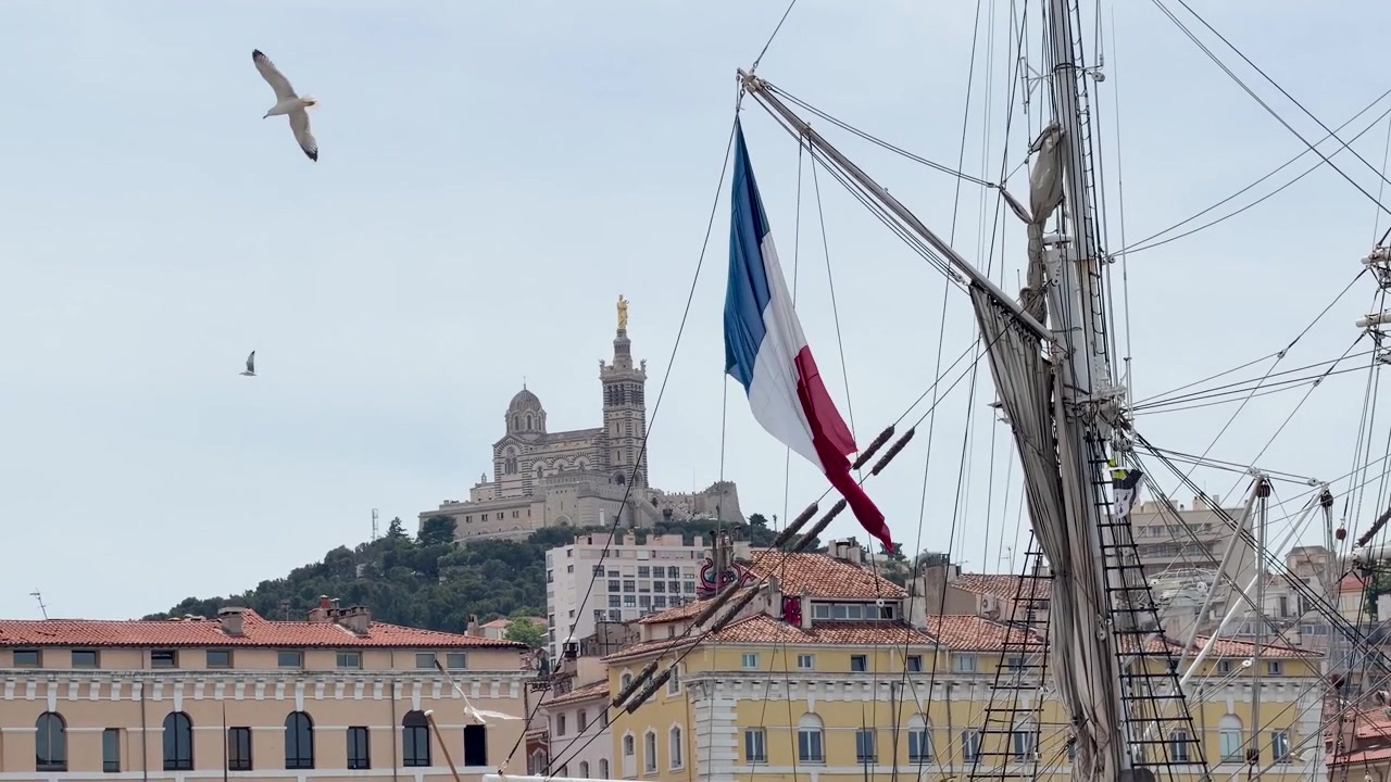 Et fransk trefarvet flag blafrer fra en sejlbådsmast med Notre-Dame de la Garde i baggrunden