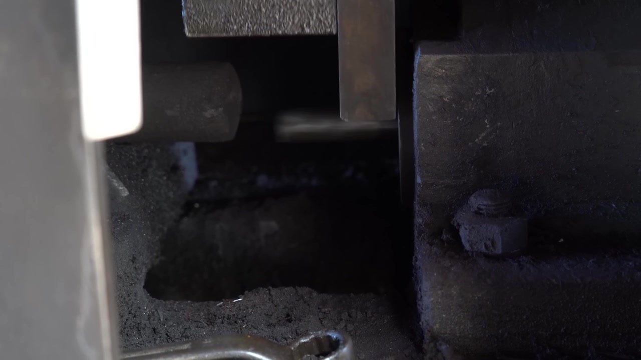 Heavy steel forging tooling inside a dim workshop with a chrome plated golf club component resting beneath the die block