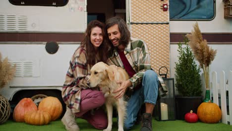 Portrait-of-a-happy-brunette-girl-and-her-boyfriend-in-a-green-plaid-shirt-petting-their-big-cream-dog-near-a-camp-trailer-during-a-picnic-outside-the-city-in-summer