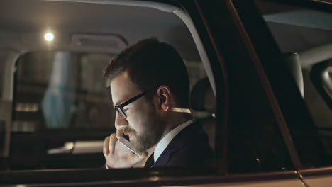 Side-View-Of-A-Man-Wearing-A-Jacket-Talking-On-The-Phone-And-His-Wife-In-The-Background,-Sitting-In-A-Moving-Car-At-Night