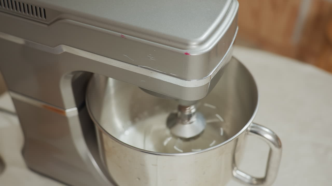 Rotational angle of electric stand mixer actively whipping white ingredients inside stainless steel bowl on kitchen table, accompanied by wooden baking tool, under warm lighting with soft background