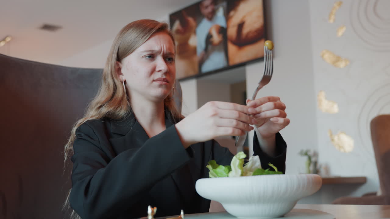 Forretningskvinde på café inspicerer salat med forvirret udtryk, opdager uventet genstand på gaffel, moderne restaurantinteriør, skål med grønt, anspændt reaktion og stille klage, nærbillede fanger