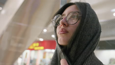 close up of lady in glasses gently holding scarf near face while standing indoors with blurred mall background, soft lighting highlighting facial features and cozy winter clothing style