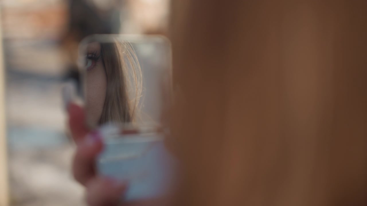 Primo piano del riflesso in uno specchietto compatto di una giovane donna che controlla trucco e unghie, lo specchietto d'argento tenuto in mano mostra uno sfondo stradale sfocato, luce solare soffusa e bokeh, un momento intimo da beauty blogger con delicati.