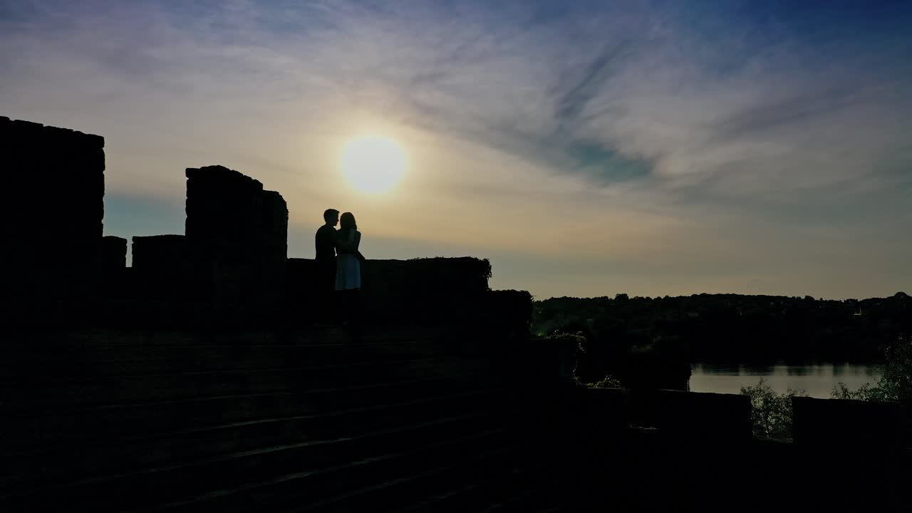 Romantic couple in love spending time together at sunset. Young man kisses girlfriend standing in a castle over the water in the evening.