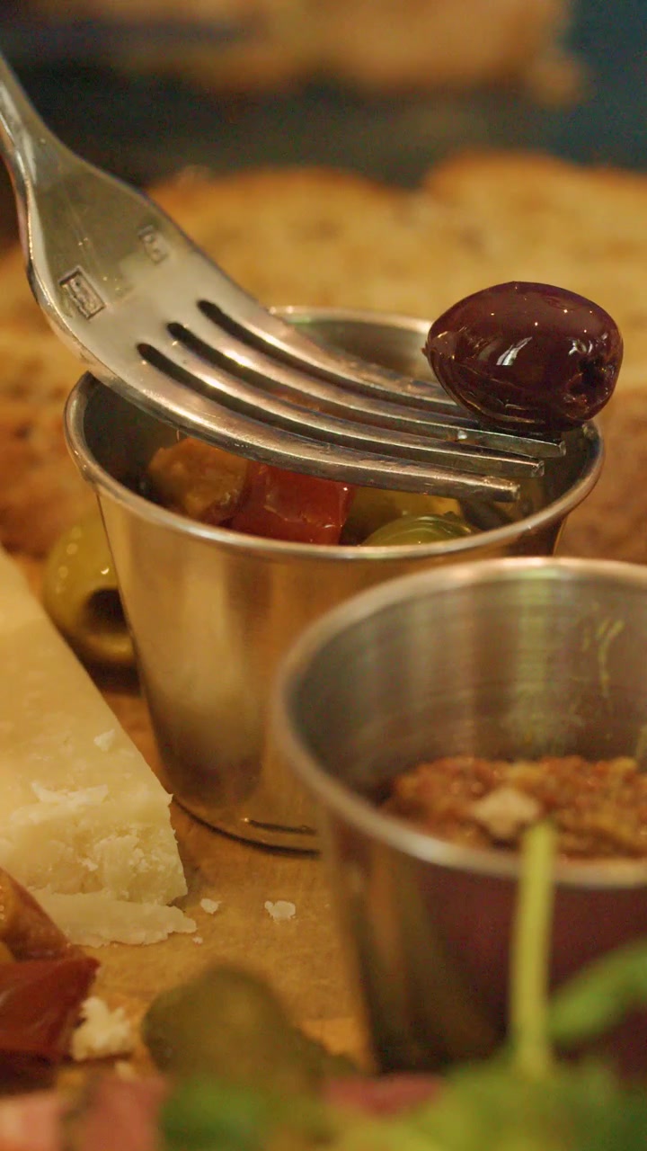 Una forchetta solleva un'oliva Kalamata da una piccola coppetta di metallo su un piatto di antipasti con formaggio, sottaceti e pane, sotto una calda illuminazione interna.