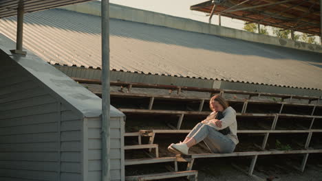 lady seated alone on stadium bleachers holding her bag in thoughtful pose, gazing into distance, background includes metal roof structure and natural setting with trees and greenery