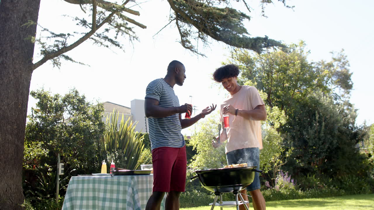 Grilling food, two multiracial male friends enjoying drinks and chatting in backyard