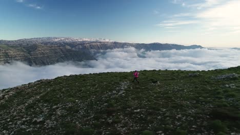 El-Hombre-Corre-Con-El-Perro-En-La-Cima-De-Una-Montaña-Rocosa-Con-Vistas-A-Las-Nubes-En-El-Valle,-Antena