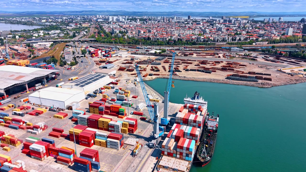 Burgas, Bulgaria, 1 May 2026: Cargo Ship Loading Near Urban Industrial Port. Aerial view of docked container ship, stacked containers, cranes, storage yard, water, and urban background.