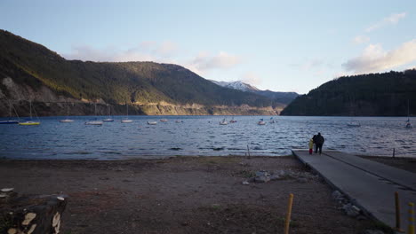 a family strolls near lake lacar, with boats on the water and mountains in the background