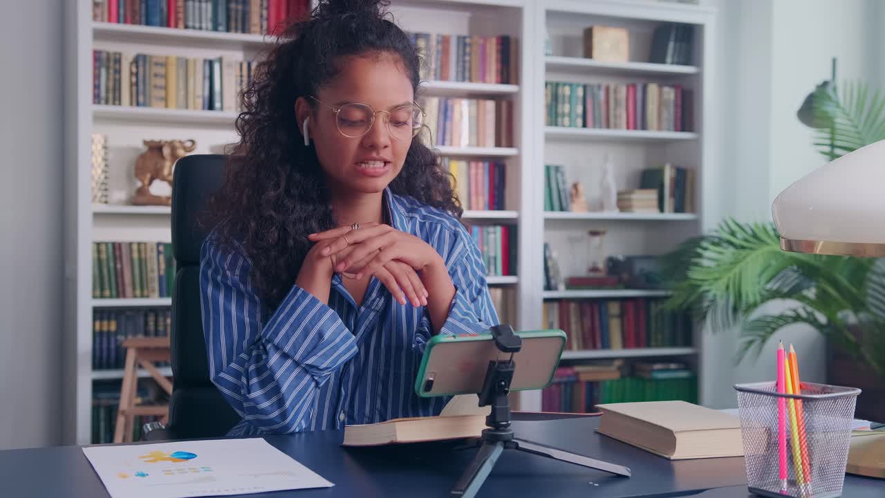 Young student indian woman sits at table reads book conducting video broadcast