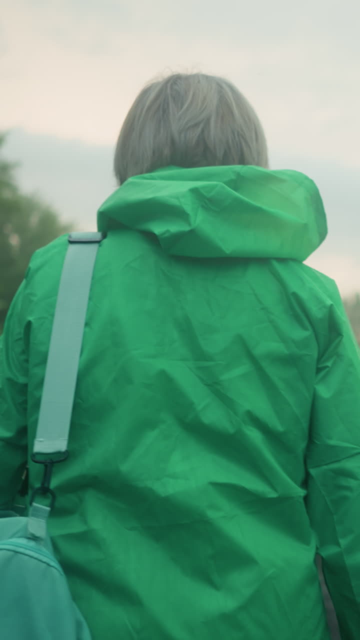 Back view of an elderly woman in a green raincoat carrying a mint-colored bag, walking along a path surrounded by greenery and trees, under a soft, cloudy sky as she moves forward