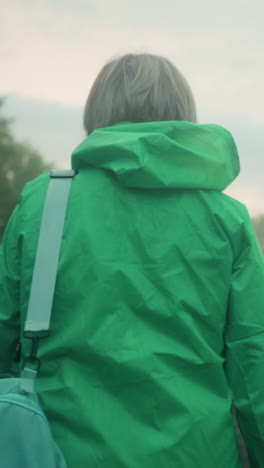 vista trasera de una mujer anciana en un impermeable verde que lleva una bolsa de color menta, caminando por un camino rodeado de vegetación y árboles, bajo un cielo suave y nublado mientras se mueve hacia adelante