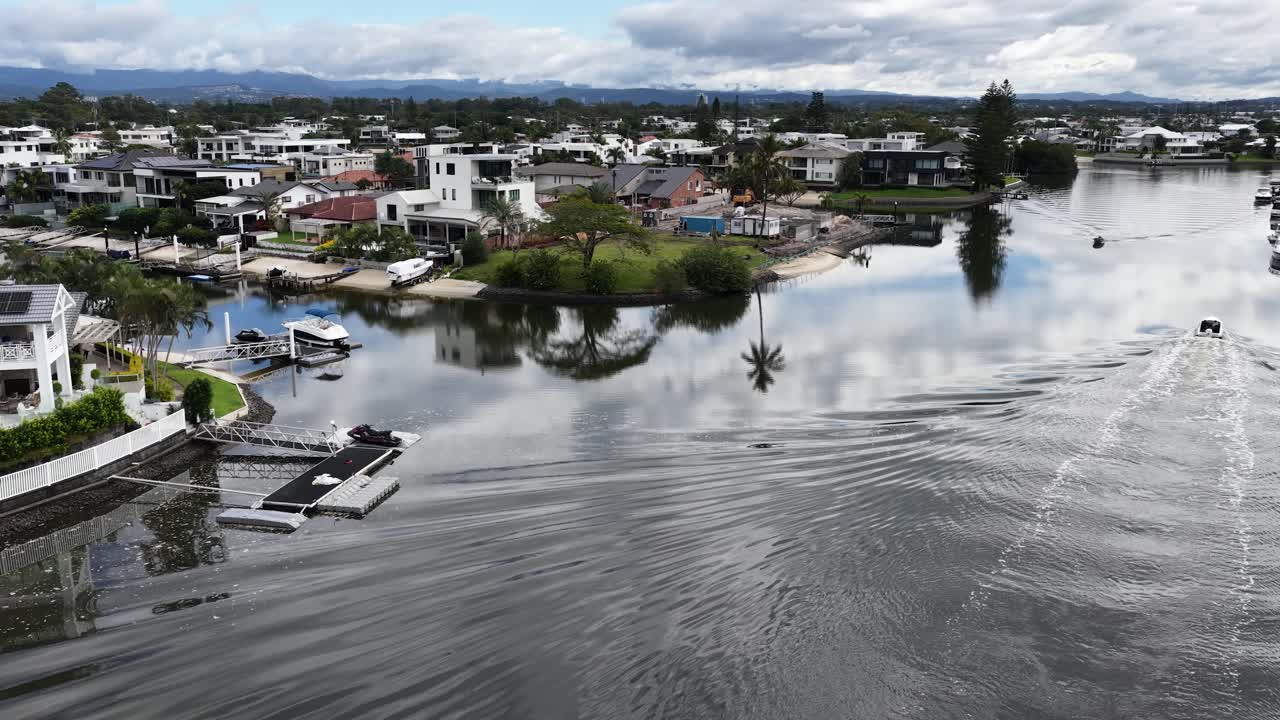 A boat navigates serene Gold Coast canals, reflecting luxury homes and lush greenery under a cloudy sky