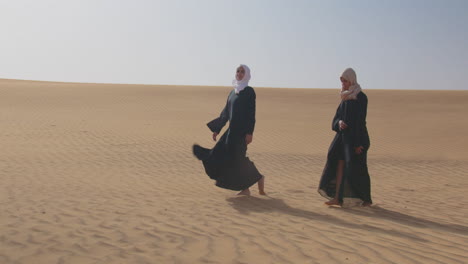 Zwei-Muslimische-Frauen,-Die-Traditionelle-Kleidung-Und-Hijab-Tragen,-Gehen-Zusammen-In-Einer-Windigen-Wüste