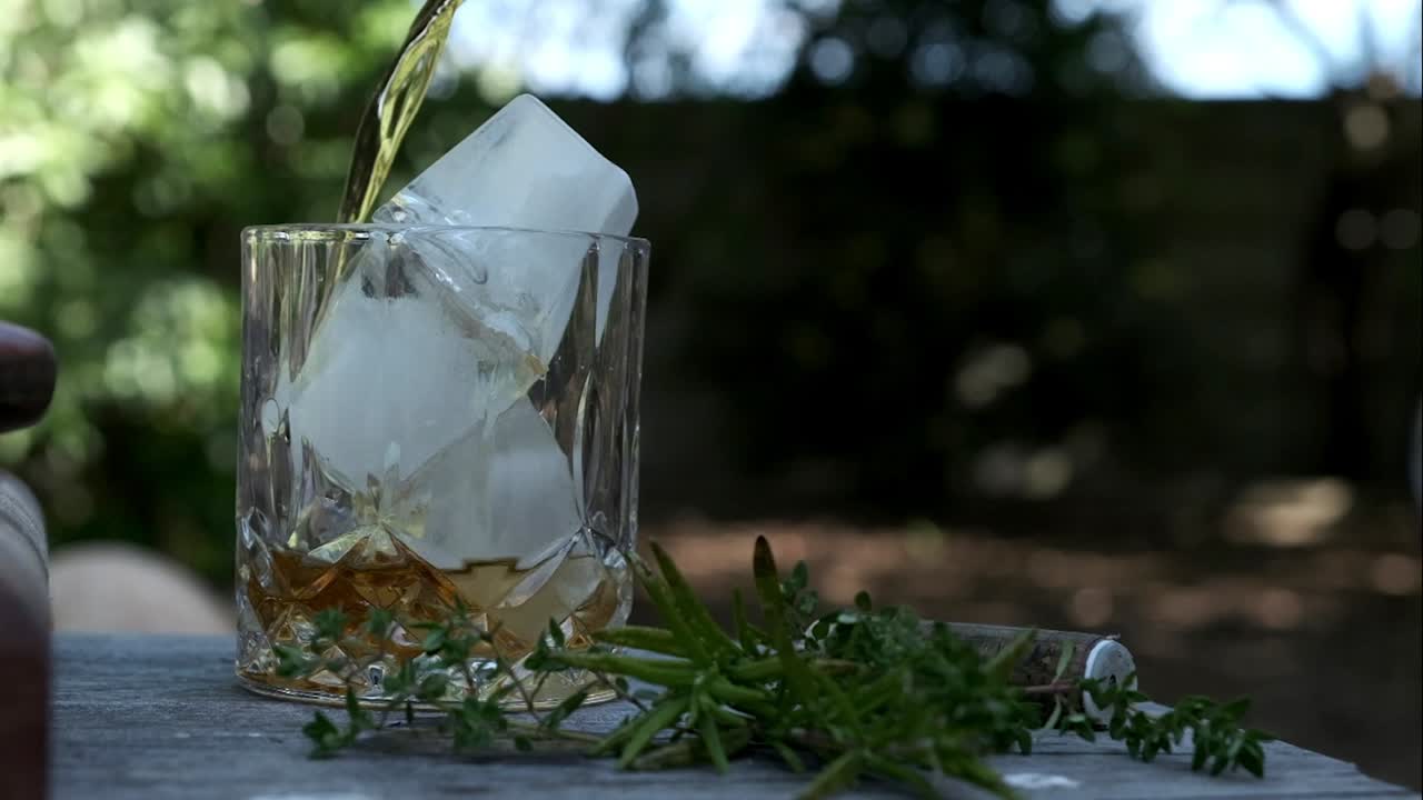 el whisky se vierte en una clase de pelota alta con hielo en un entorno al aire libre bajo algunos árboles