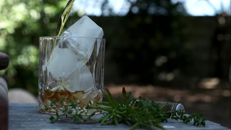 whiskey is poured into a high ball class with ice in an outdoor setting under some trees