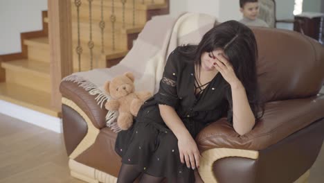 beautiful middle eastern mother with migraine sitting on the armchair and her two little children running around