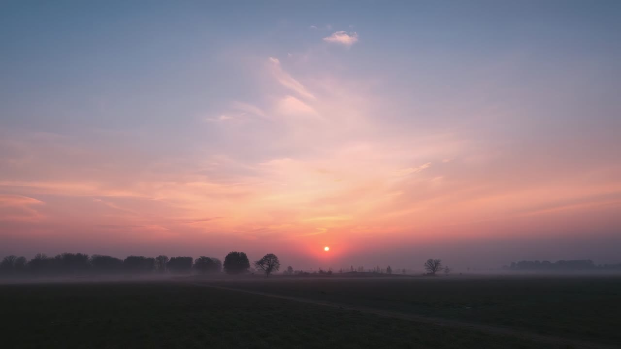Rising sun casting warm light across farmland at dawn, with misty tree line welcoming new day